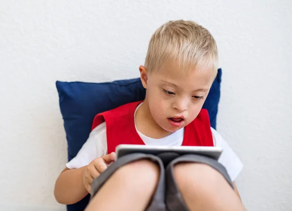 Ett porträtt av Down-syndromet School Boy sitter på golvet, med hjälp av Tablet. — Stockfoto