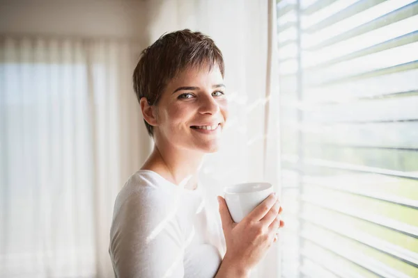 Retrato de mujer joven con café de pie junto a la ventana en el interior de casa . —  Fotos de Stock