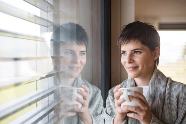 Portrait of young woman with coffee standing indoors by window at home. — Stock Photo, Image