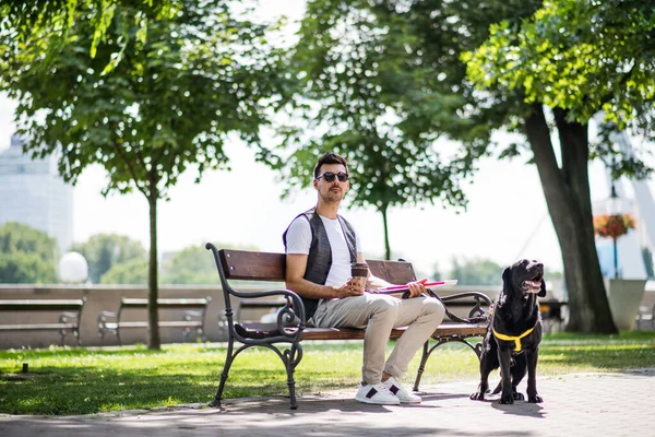 Young blind man with white cane and guide dog sitting in park in city. — Stock Photo, Image