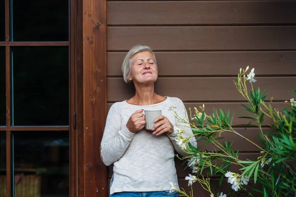 Eine Seniorin mit Kaffee, die im Sommer draußen auf einer Terrasse steht. — Stockfoto