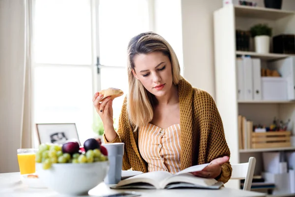 Jonge vrouw die thuis aan tafel zit, leest en eet. — Stockfoto