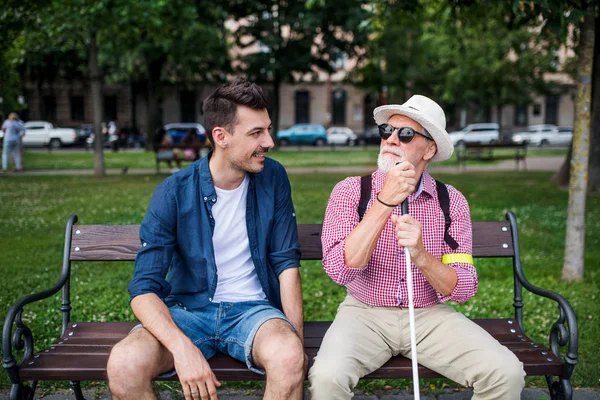Молодой человек и слепой старший с белой тростью сидит на скамейке в парке в городе . — стоковое фото