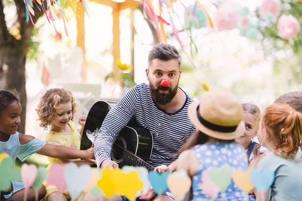 Homme avec de jeunes enfants au sol à l'extérieur dans le jardin en été, jouer de la guitare . — Photo