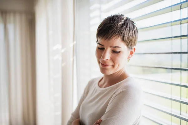 Portrait de jeune femme debout par la fenêtre à l'intérieur à la maison, rêvant . — Photo