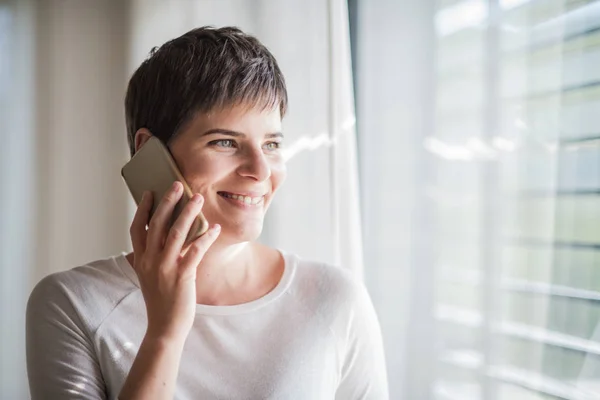 Akıllı telefonlu genç bir kadın evin penceresinin yanında duruyor.. — Stok fotoğraf