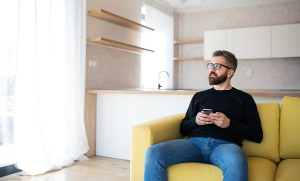 Happy mature man sitting on sofa in unfurnished house, holding smartphone. — Stock Photo, Image