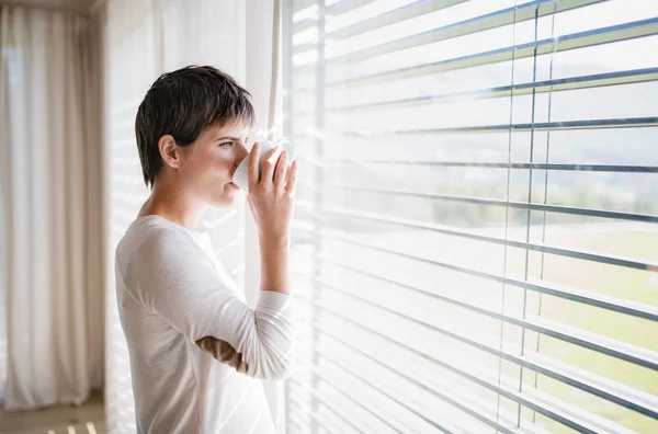 Retrato de jovem mulher com café em pé à janela dentro de casa . — Fotografia de Stock