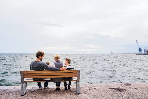 Rear view of young family with two small children on bench outdoors on beach. — Stockfoto