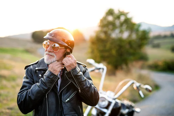 Um homem idoso viajante com moto no campo, vestindo capacete . — Fotografia de Stock