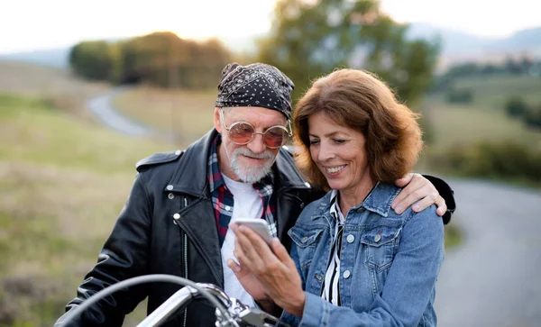 Una pareja de viajeros mayores con moto en el campo, utilizando el teléfono inteligente . —  Fotos de Stock