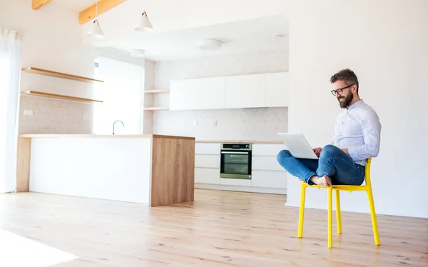 Un homme mature assis sur une chaise dans une nouvelle maison non meublée, à l'aide d'un ordinateur portable . — Photo