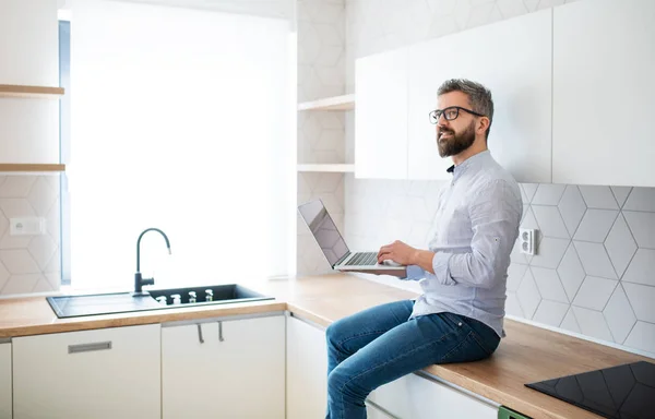 Un homme mature avec ordinateur portable assis dans la cuisine dans une maison neuve non meublée . — Photo