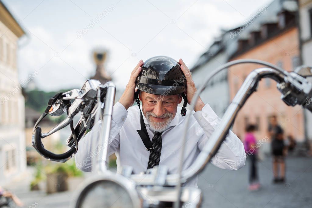 A senior businessman with motorbike in town, putting on helmet.