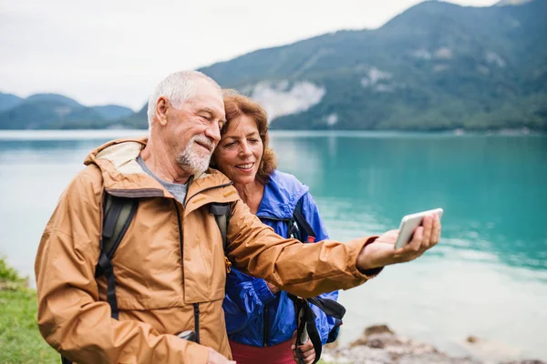 Starší důchodce pár stojící u jezera v přírodě, přičemž selfie. — Stock fotografie