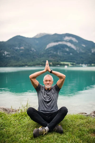 Старший чоловік пенсіонер, сидячи на озері в природі, займається вправами йоги . — стокове фото