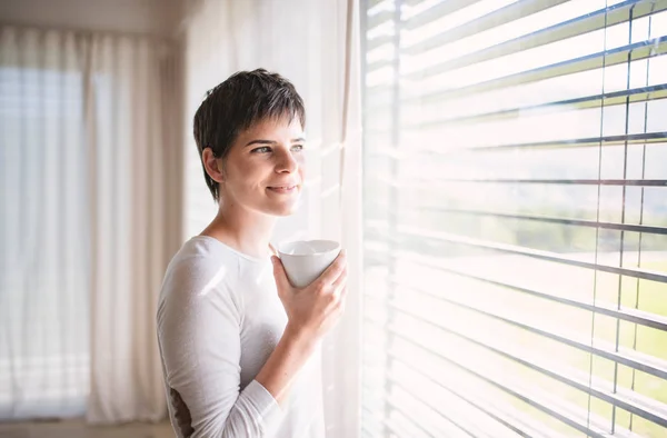 Portret młodej kobiety z kawą stojącej przy oknie w domu. — Zdjęcie stockowe