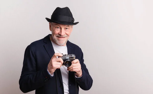 Portret van een oudere man met hoed en camera in een studio. — Stockfoto