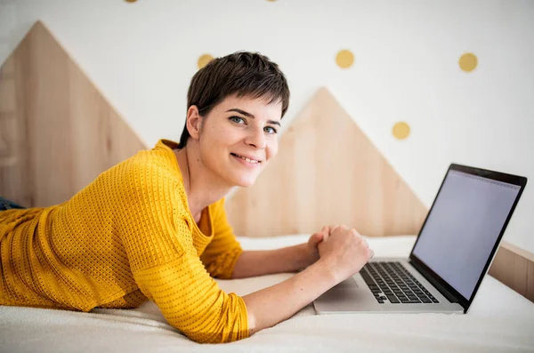 Young woman with laptop lying on bed in bedroom indoors at home. — Stock Photo, Image