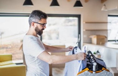 Kutuları ve elbise askısı olan olgun bir adam yeni mobilyasız eve taşınıyor..