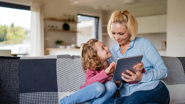 Una linda niña pequeña con madre en el sofá en el interior de casa, utilizando la tableta . — Foto de Stock