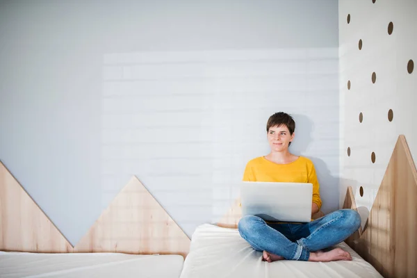 Wanita muda dengan laptop duduk di tempat tidur di kamar tidur di dalam rumah . — Stok Foto
