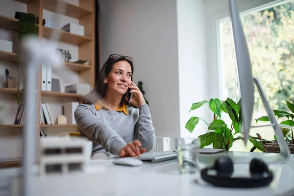 Empresária atraente sentada na mesa dentro de casa no escritório, usando smartphone . — Fotografia de Stock