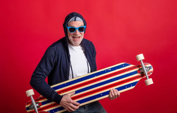 En senior man med longboard i en studio på röd bakgrund, tittar på kameran. — Stockfoto