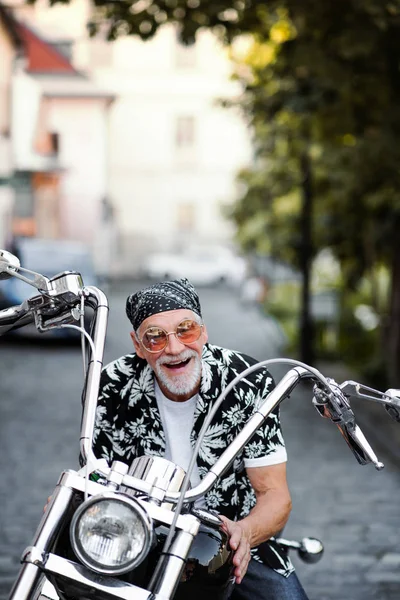 A front view of cheerful senior man traveller with motorbike in town. — Stock Photo, Image