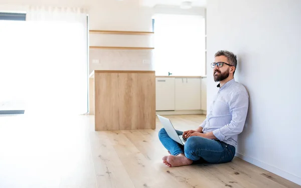 Un homme mature assis sur le sol dans une nouvelle maison non meublée, à l'aide d'un ordinateur portable . — Photo