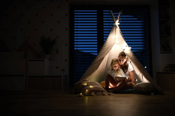 Junge Mutter mit Tochter sitzt drinnen im Schlafzimmer und liest ein Buch. — Stockfoto