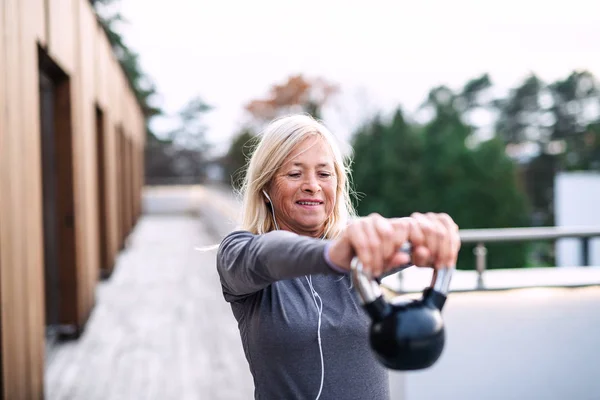 Una donna anziana con auricolari e kettlebell all'aperto facendo esercizio . — Foto Stock