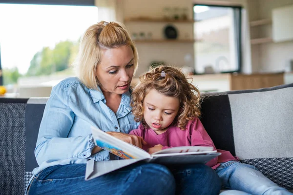 Una piccola ragazza carina con la madre sul divano al chiuso a casa, libro di lettura . — Foto Stock