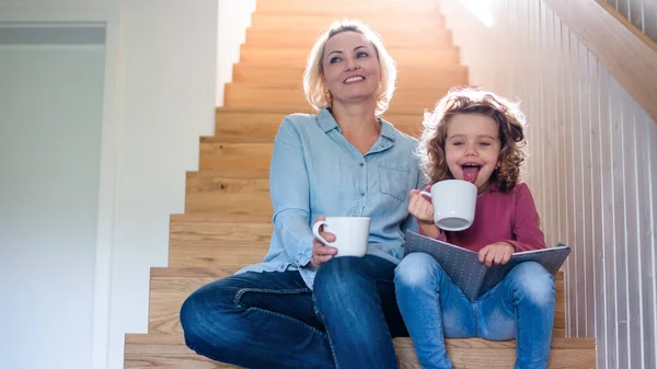 Uma menina pequena bonito com a mãe dentro de casa, lendo na escada . — Fotografia de Stock