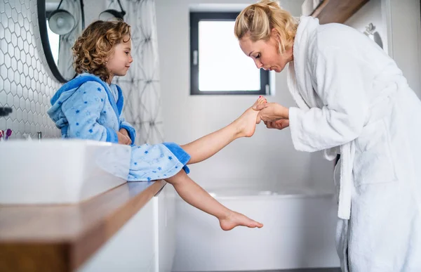 Une jolie petite fille avec mère dans la salle de bain à l'intérieur à la maison, peindre des ongles . — Photo