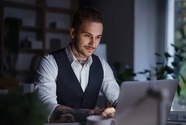 Um homem de negócios com laptop sentado na mesa dentro do escritório, trabalhando até tarde . — Fotografia de Stock