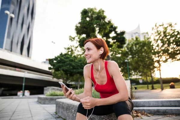 スマートフォンを使って市内のイヤホンを持つ若い女性ランナー. — ストック写真
