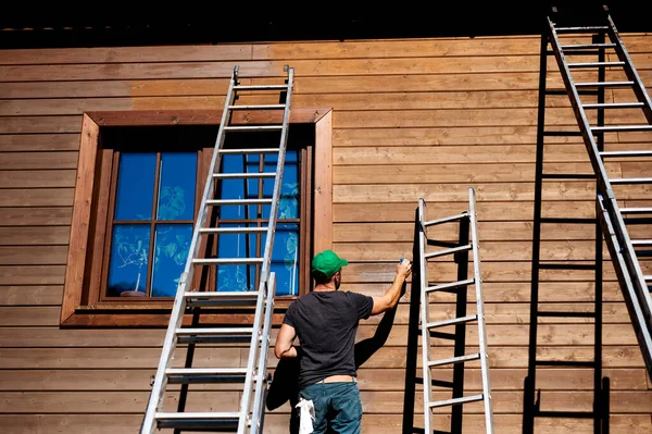 Um homem maduro ao ar livre no verão, pintando casa de madeira . — Fotografia de Stock