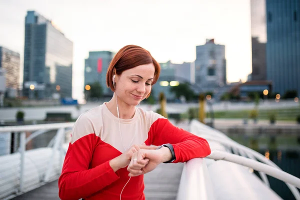 スマートウォッチを使用して、市内のイヤホンを持つ若い女性ランナー. — ストック写真