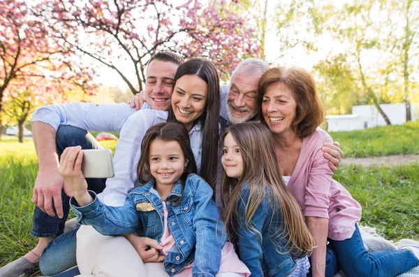 Trzy pokolenia rodziny siedzą na zewnątrz w wiosennej przyrodzie, robiąc selfie. — Zdjęcie stockowe