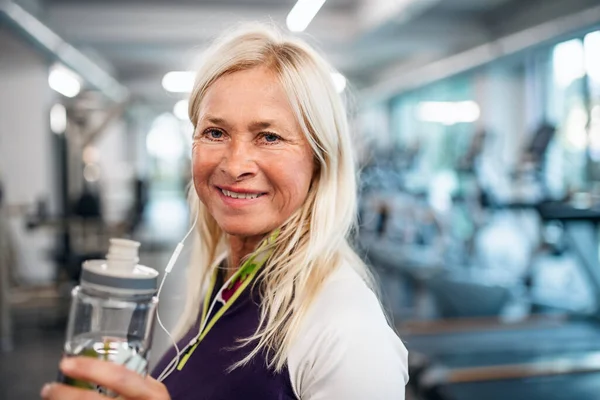 Une femme âgée avec écouteurs et bouteille d'eau debout dans la salle de gym . — Photo
