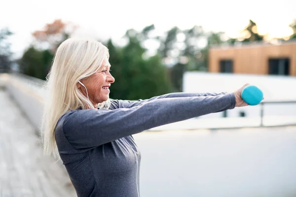 Starší žena se sluchátky a činkami venku cvičí. — Stock fotografie