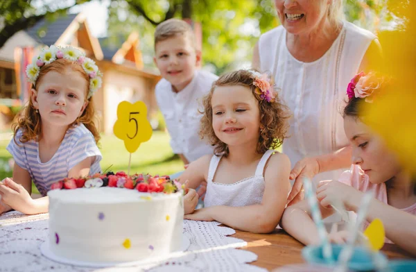 Dzieci z ciastem stojące wokół stołu na przyjęciu urodzinowym w ogrodzie latem. — Zdjęcie stockowe