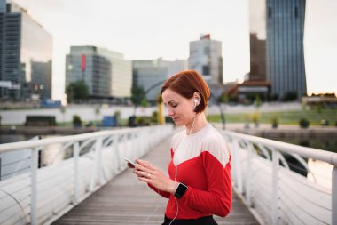Şehirde kulaklıklı genç kadın koşucu, akıllı telefon kullanarak.