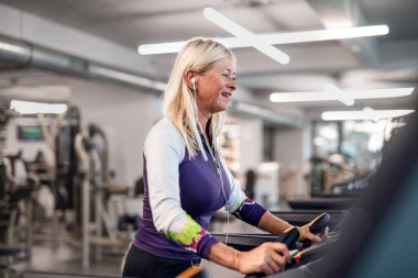 Spor salonunda kulaklıkları olan, kardiyo çalışması yapan son sınıf öğrencisi bir kadın..
