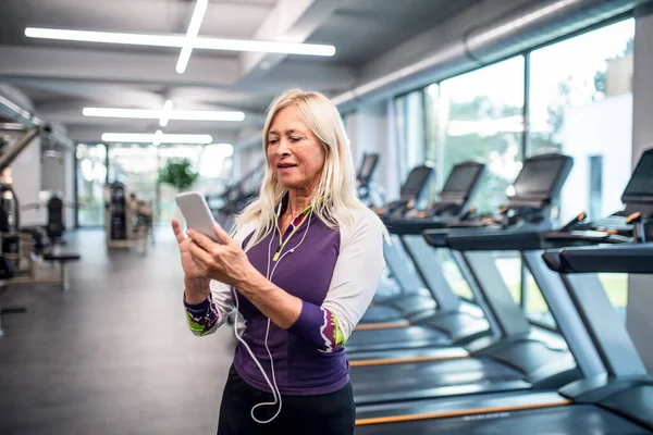 Een oudere vrouw met smartphone in de sportschool rusten na het doen van oefening. — Stockfoto