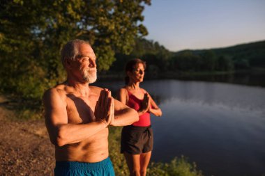Senior couple in swimsuit standing by lake outdoors doing yoga.
