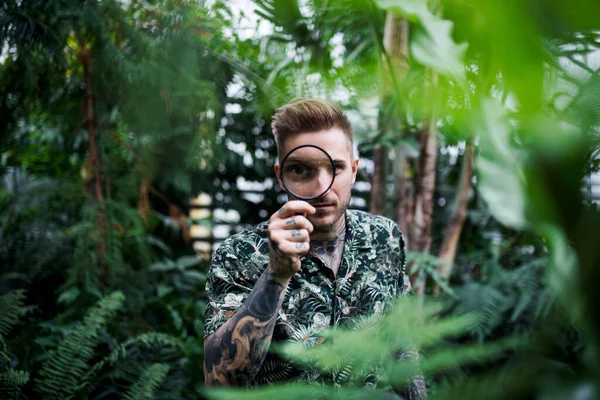 Young man with magnifying glass standing in botanical garden. — Stock Photo, Image