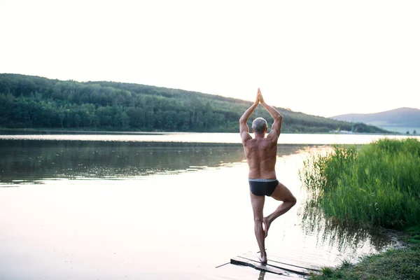 Rear view of senior man in swimsuit standing by lake outdoors doing yoga. — Stockfoto