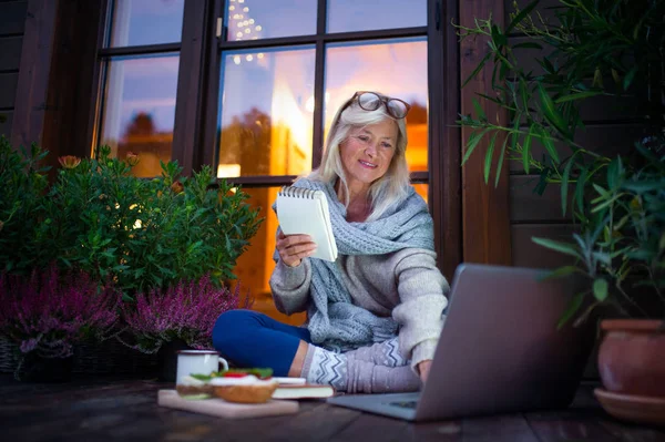 Senior woman with laptop sitting outdoors on terrace, working in the evening. — Stockfoto